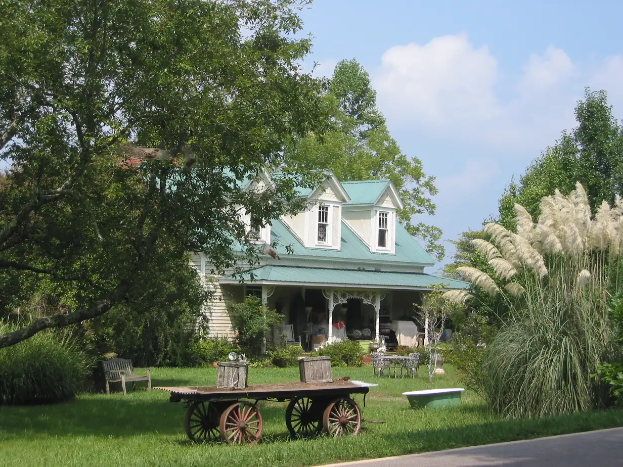 Two-story farmhouse with teal metal roof and dormers, wide front porch, old wooden wagon and pampas grass on the lawn
