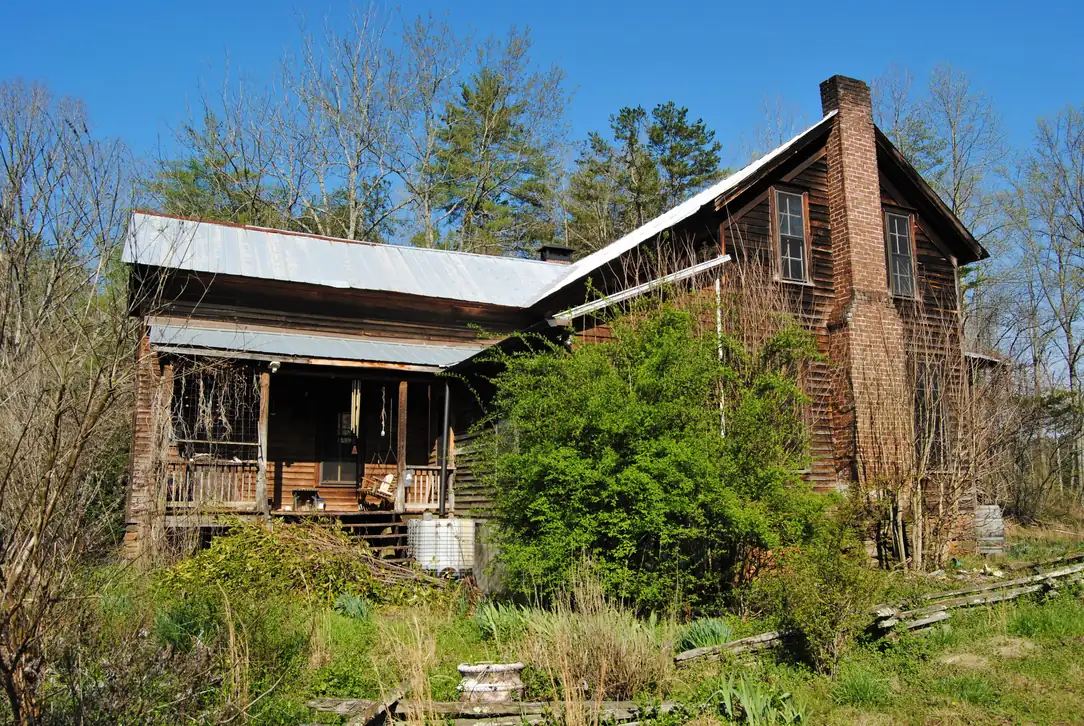 Weathered wooden farmhouse with metal roof, brick chimney and front porch in an overgrown spring yard