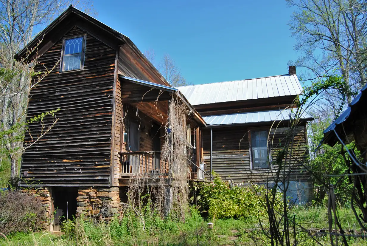 Weathered wooden farmhouse on a stacked-stone foundation with a vine-draped side porch under blue sky