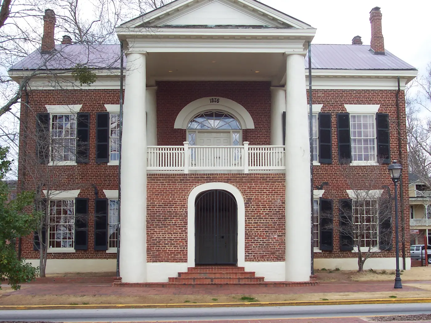 Red-brick two-story building with tall white columns, black shutters and an 1836 fanlight over the balcony