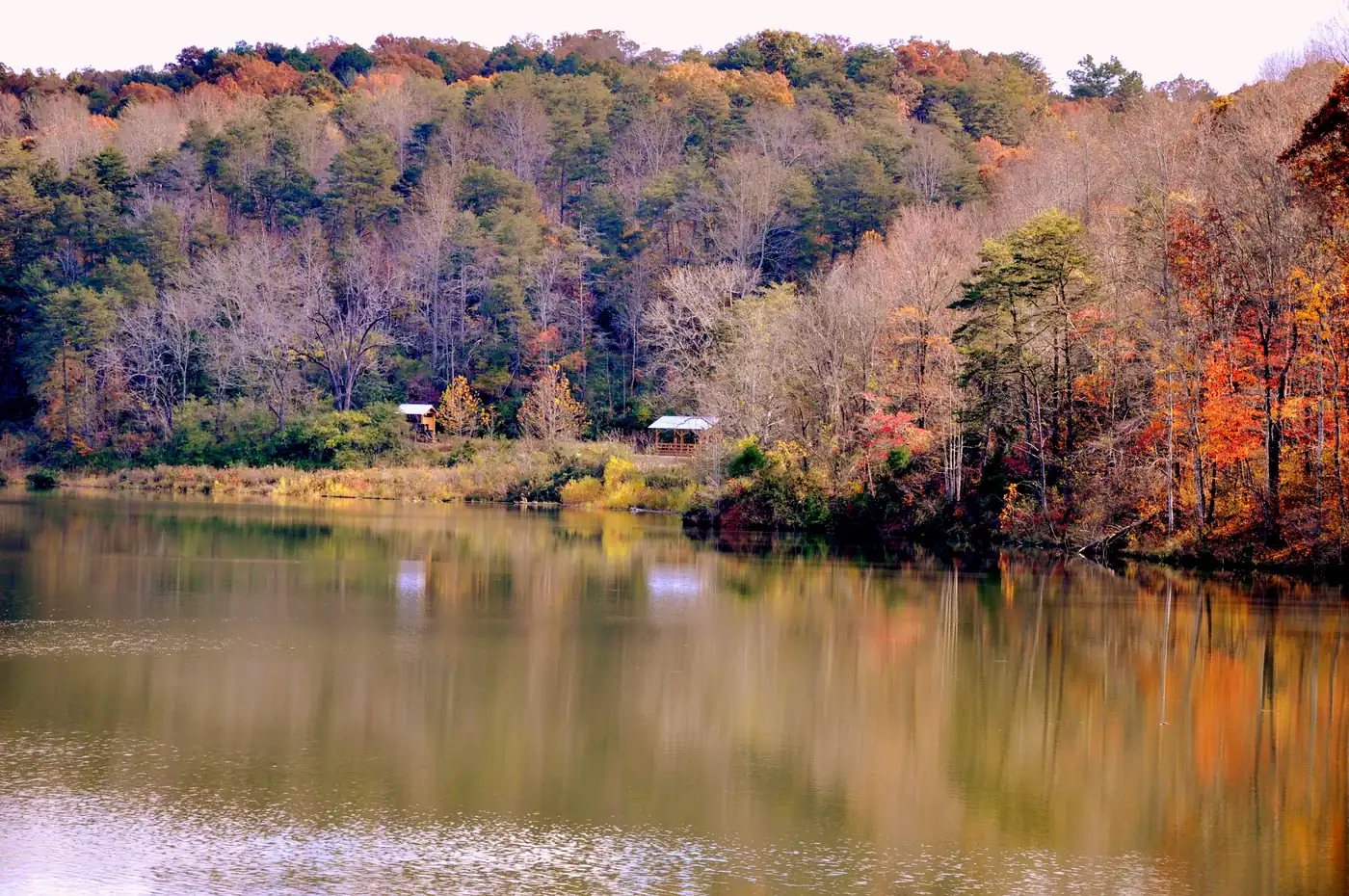 Calm lake reflecting a hillside of orange, red and gold autumn trees with two small shoreline shelters