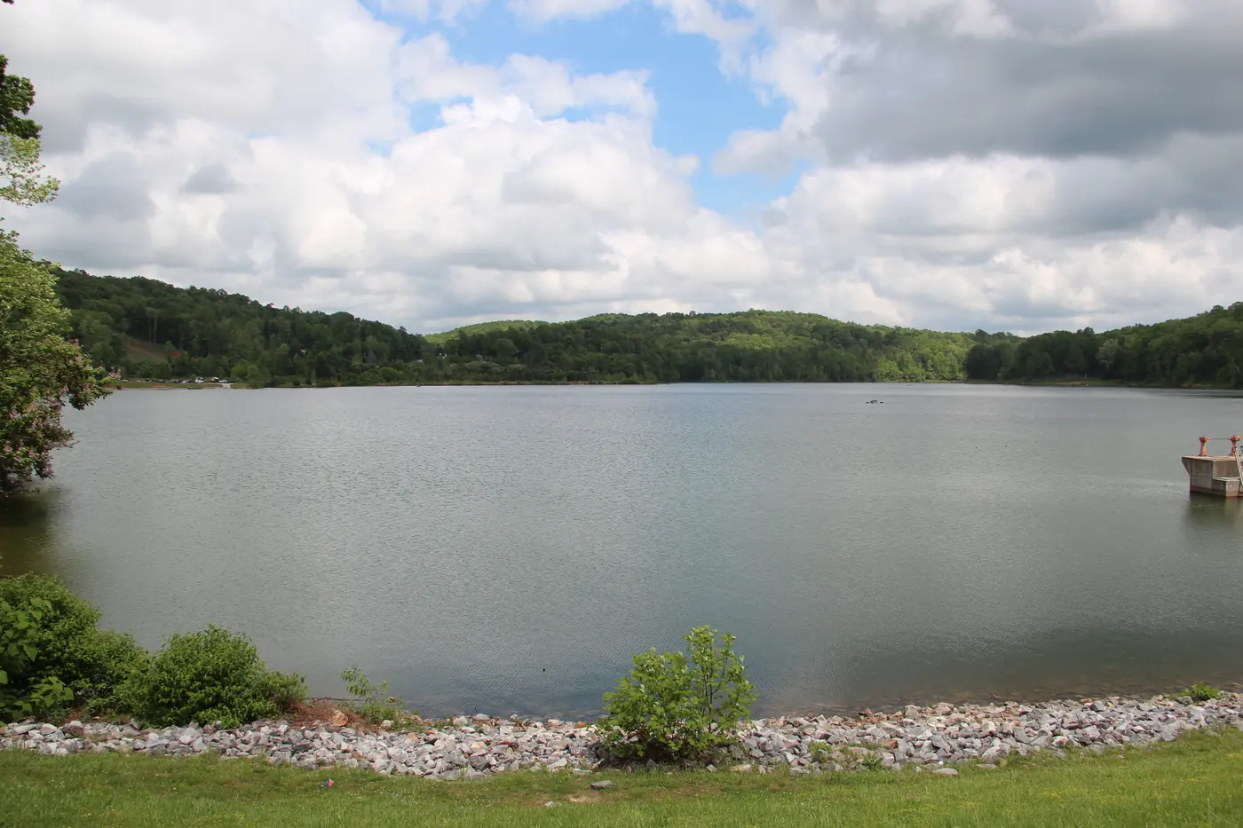 Wide lake ringed by green wooded hills under big white clouds, with a stone-lined grassy shore in front