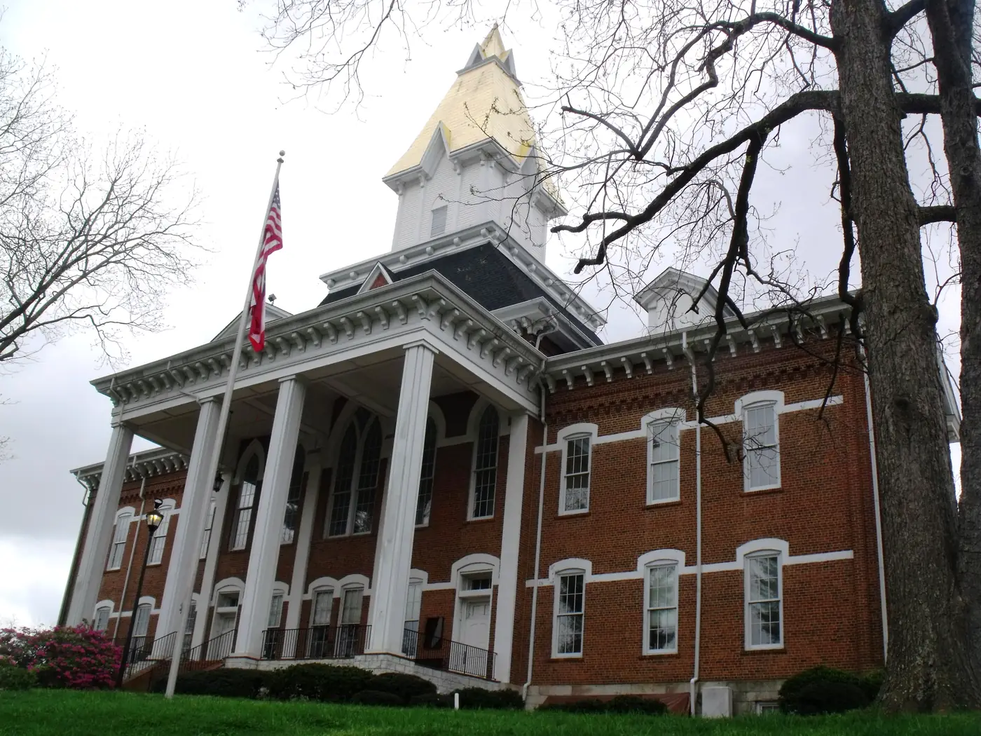 Red-brick building with white columns and a gold-leaf spire, American flag and pink azaleas in front