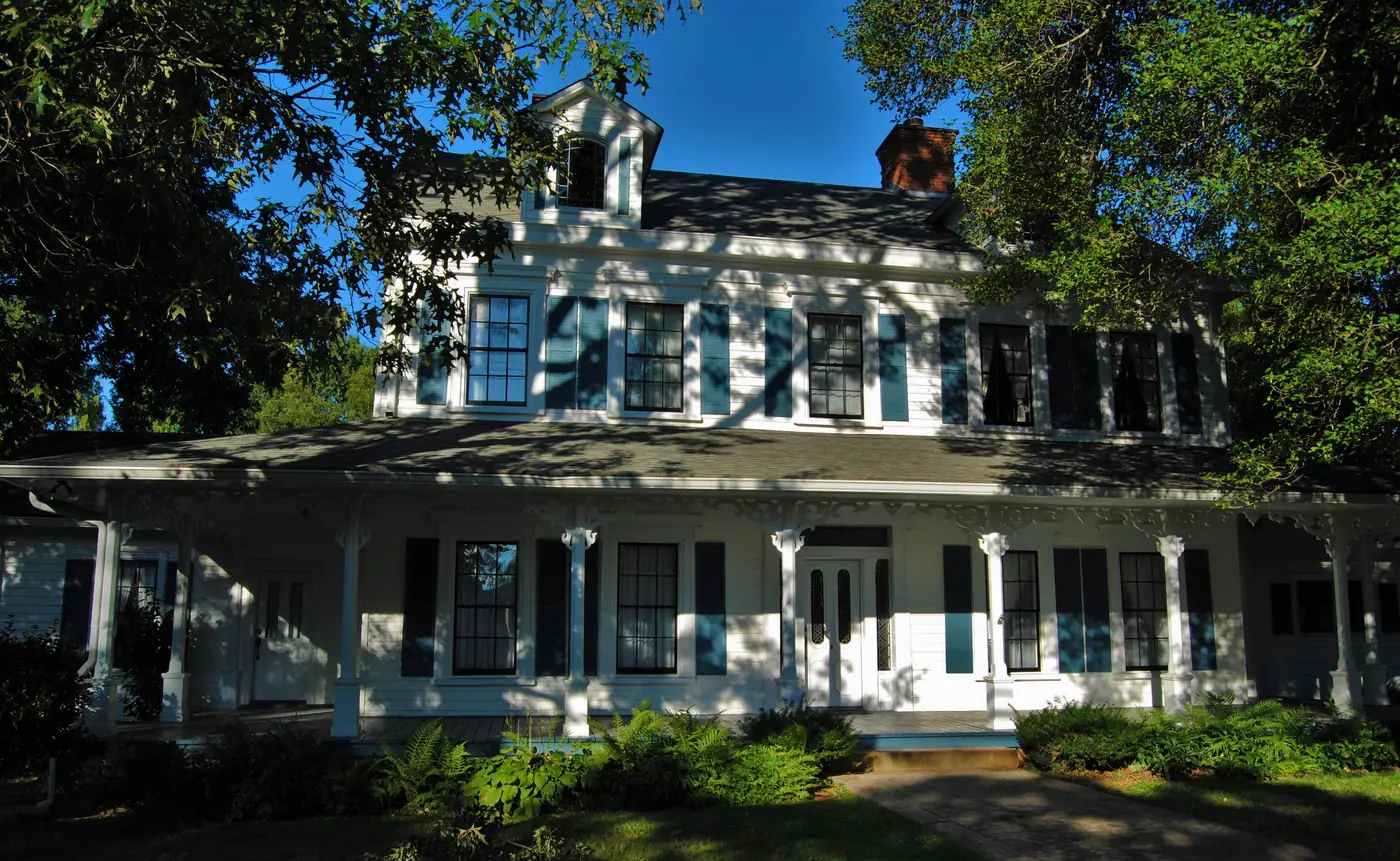 White two-story house with blue shutters and a long bracketed front porch, framed by shade trees and ferns