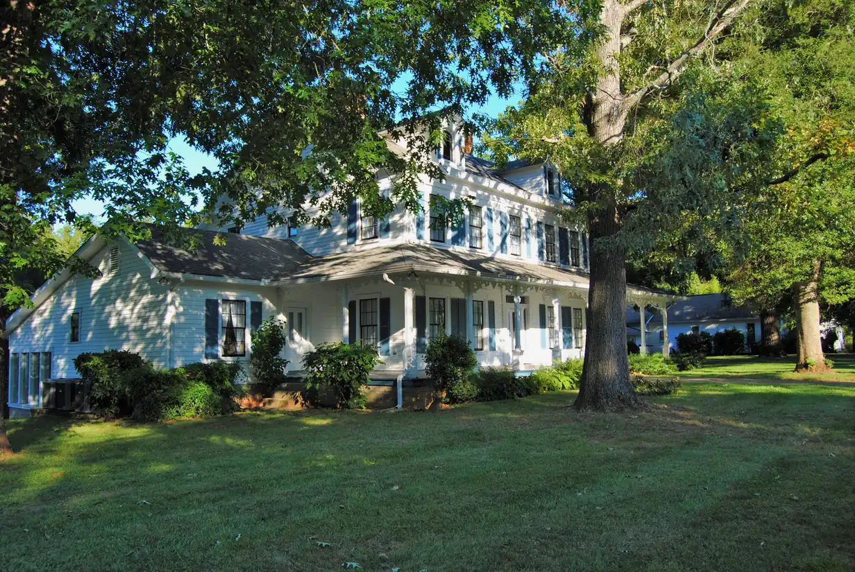Large white historic house with a wraparound porch on a wide green lawn under tall oak trees