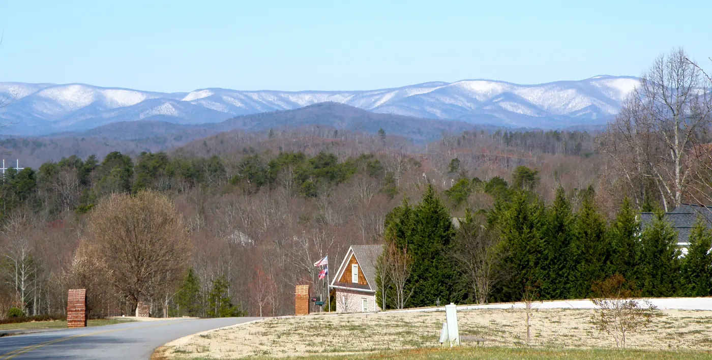 Curving road past brick entrance pillars and a house, wooded hills and snow-capped mountain ridges in the distance