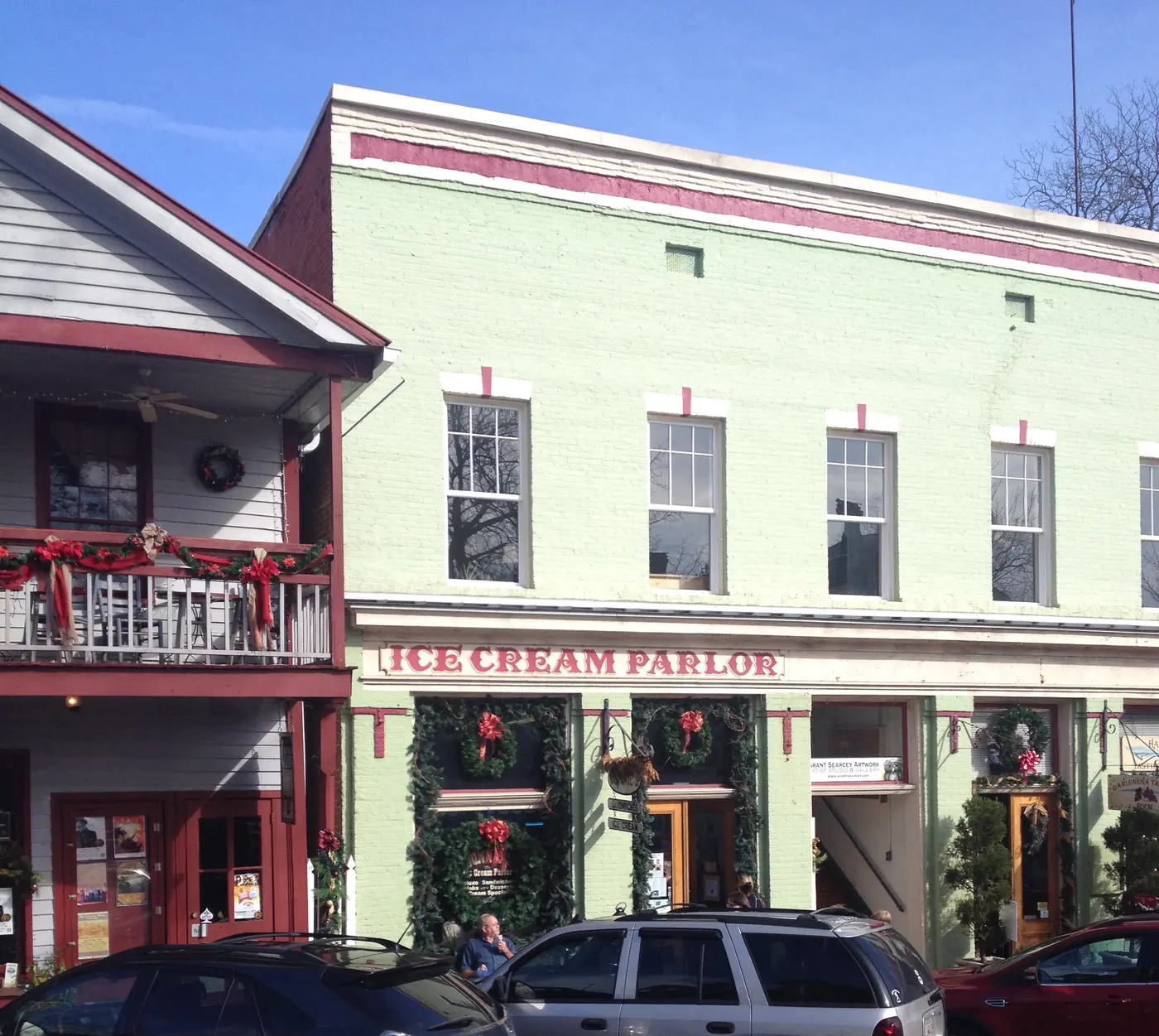 Pale green two-story brick storefront with an Ice Cream Parlor sign and holiday wreaths, cars parked in front