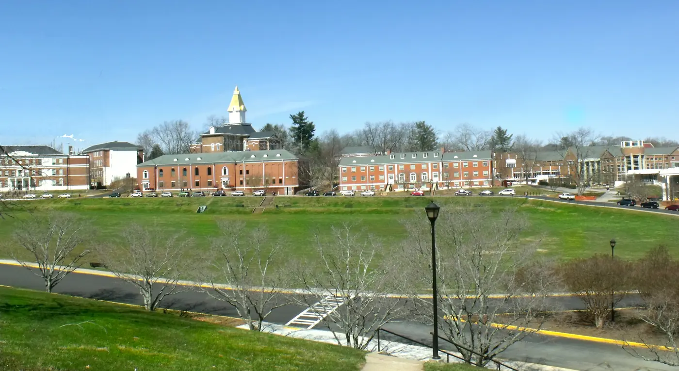 Open green field in front of red-brick campus buildings with a gold-tipped white spire, under a clear blue sky