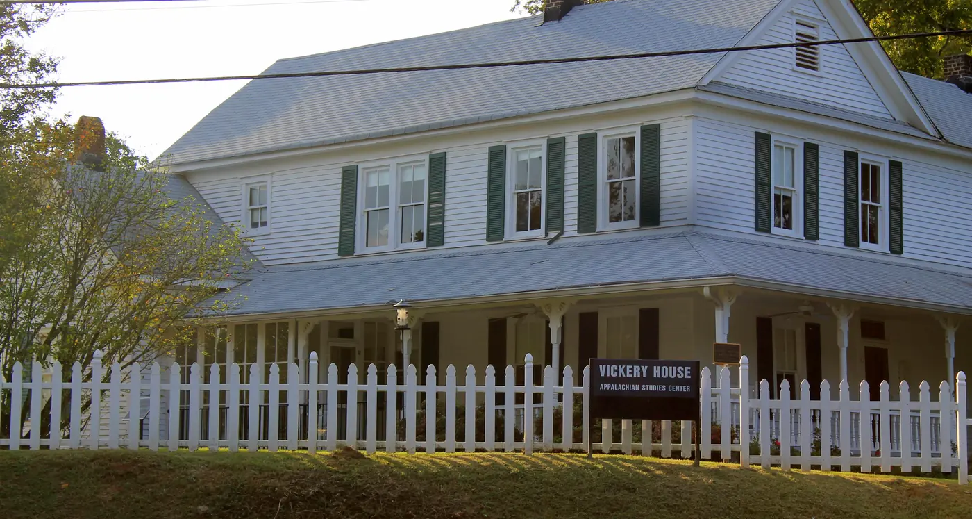 White two-story clapboard house with green shutters and a wraparound porch behind a white picket fence