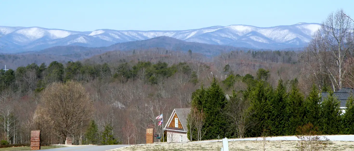 A curving neighborhood road past brick entrance pillars and a house, with wooded hills and snow-dusted mountain ridges beyond.